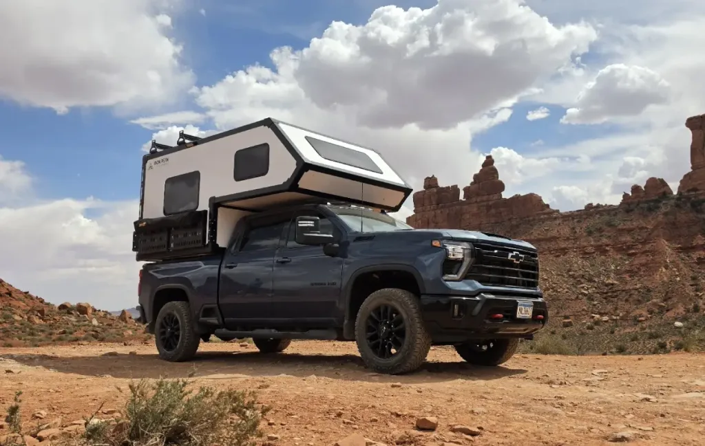 A blue pickup truck with a white camper attached is parked on a dirt road in a rocky desert landscape, with dramatic rock formations and a partly cloudy sky in the background.
