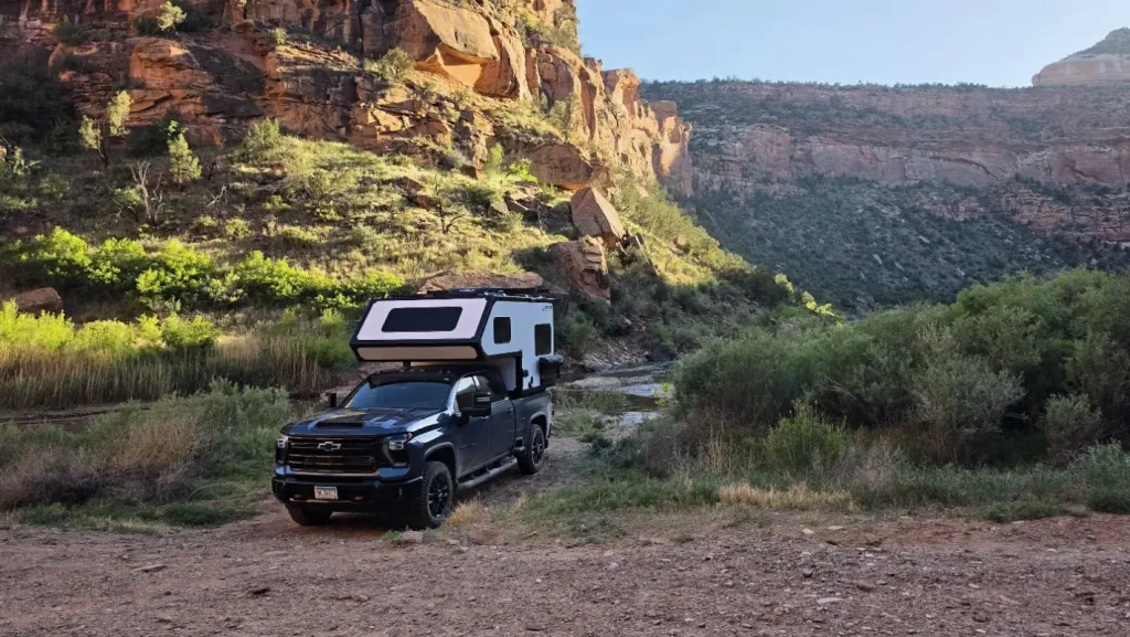 A black pickup truck with a white camper is parked on a dirt clearing surrounded by green shrubs and rocky cliffs under a clear blue sky.