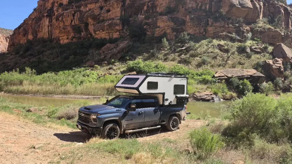 A black pickup truck with a white camper attached is parked on a dirt path beside a river, surrounded by green shrubs and tall red canyon cliffs.