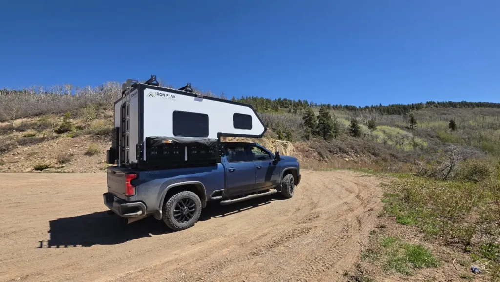 A blue pickup truck with a white pop-up camper is parked on a dirt road surrounded by green hills, sparse trees, and a clear blue sky.