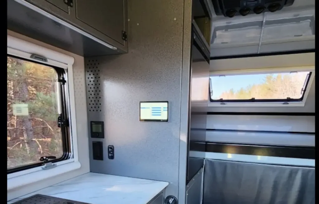 Interior of a modern camper van with a window showing trees outside, a marble countertop, gray cabinets, and a touchscreen control panel mounted on the wall. Natural light brightens the space.