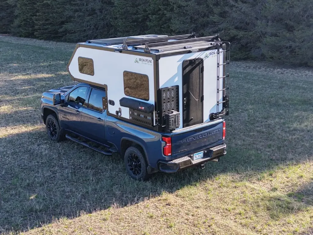A blue pickup truck with a white camper installed in its bed is parked on a grassy field near trees. The camper has windows, a ladder, and gear mounted on the back.