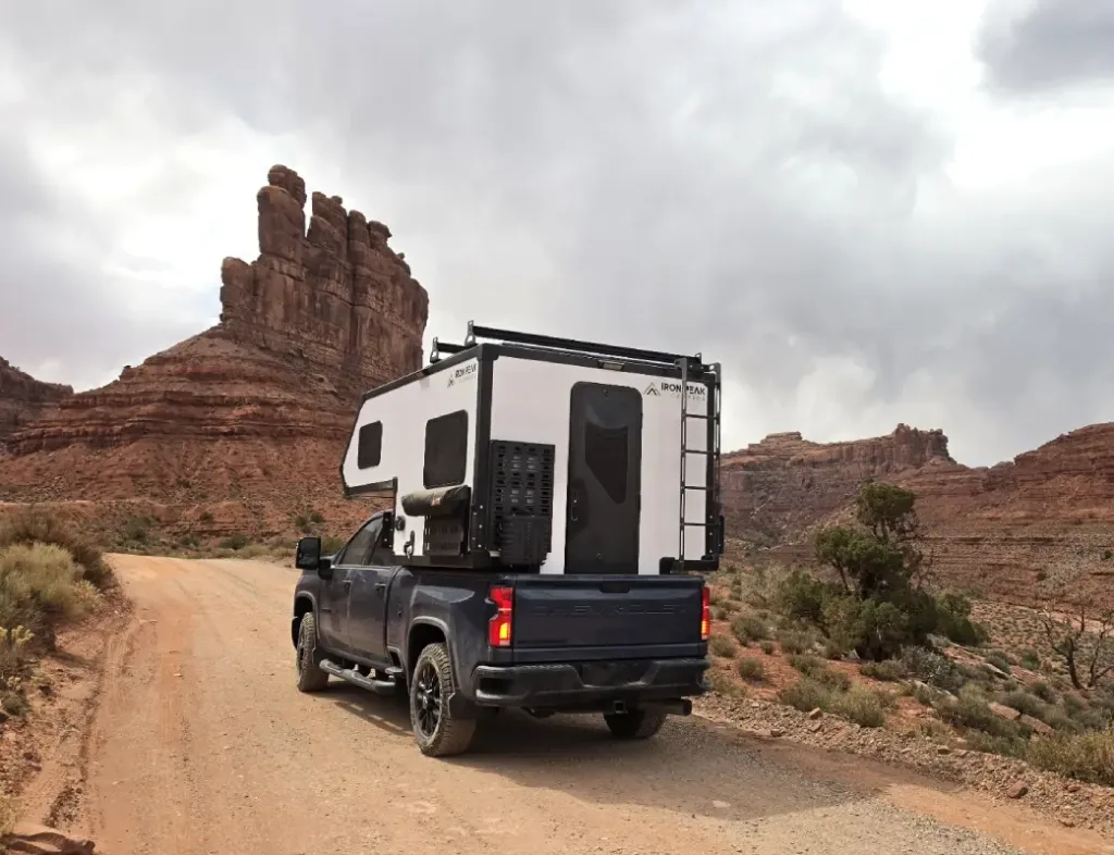 Pickup truck with a white camper shell attached on a dirt desert road, red rock formations in the background.