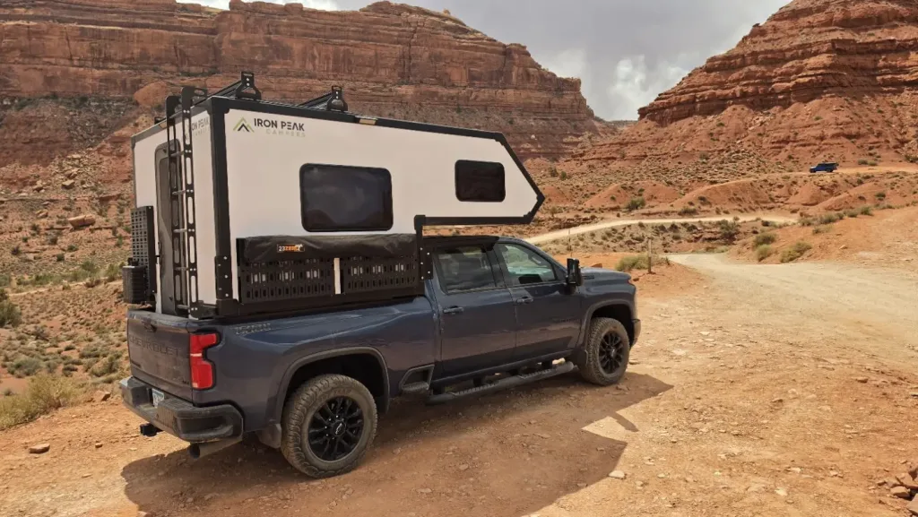 A dark blue pickup truck with a white Iron Peak camper mounted in the bed is parked on a dirt road in a rocky, red desert canyon landscape under cloudy skies.
