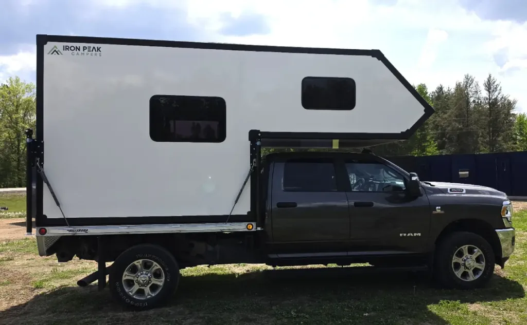 A black pickup truck with a large, white Iron Pax Campers camper attached to its bed is parked on grass, with trees and a blue container in the background under a partly cloudy sky.