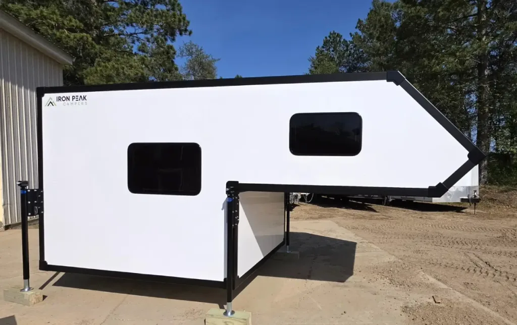 A white truck camper with black trim and two dark-tinted windows stands on support legs in a dirt area, surrounded by trees and a metal building under a clear blue sky.