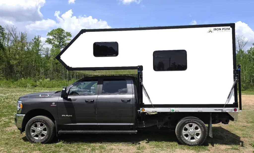 A black pickup truck with a large, white camper mounted on the back is parked outdoors on a grassy area near trees under a partly cloudy sky.