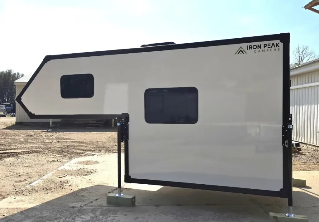 A white Iron Peak Campers truck camper sits on metal legs in a sunlit outdoor area, with two tinted windows and a logo on the upper right corner. Buildings and trees are visible in the background.