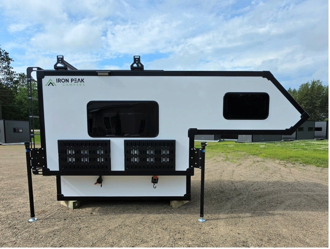 A white Iron Peak Campers truck camper with black trim is parked on gravel, with grassy areas and trees in the background under a partly cloudy sky.