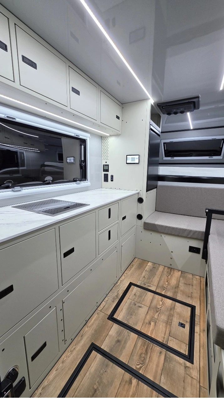Modern camper van interior with sleek white cabinets, a countertop with a sink, built-in storage drawers, wood-look flooring, and cushioned seating by large windows. LED strip lighting on the ceiling brightens the space.