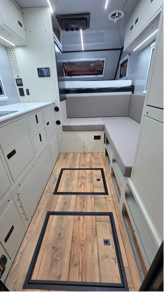 Modern camper van interior with light cabinetry, built-in sofa, large windows, overhead storage, and wood-style flooring with two large access panels. The space is tidy, well-lit, and designed for maximum functionality.