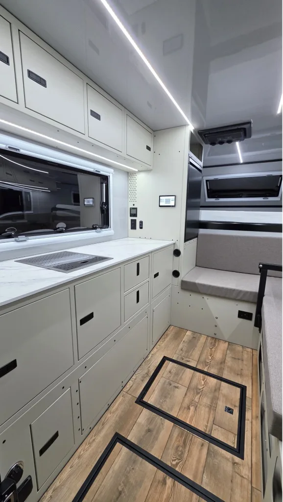 Modern camper van interior with light cabinetry, wood-look flooring, a built-in bench with gray cushion, underfloor storage, a window above the sink, and overhead LED lighting. Clean, minimalist design.