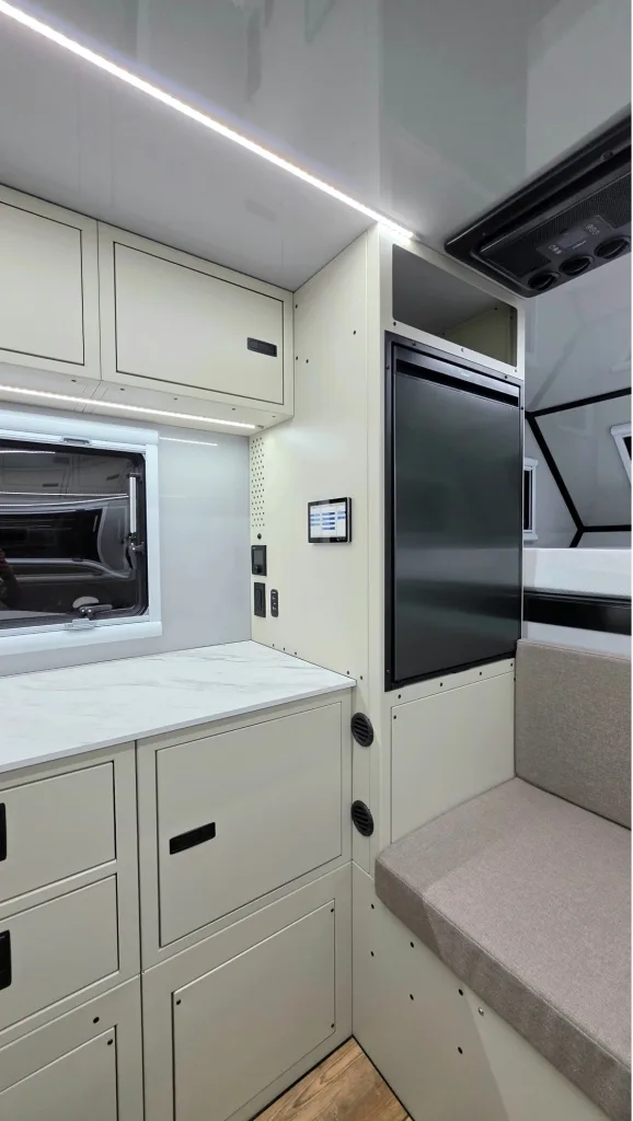 Modern RV interior featuring light-colored cabinetry, a marble-effect countertop, a large refrigerator, a window, built-in drawers, and a cushioned bench seat with overhead lighting.