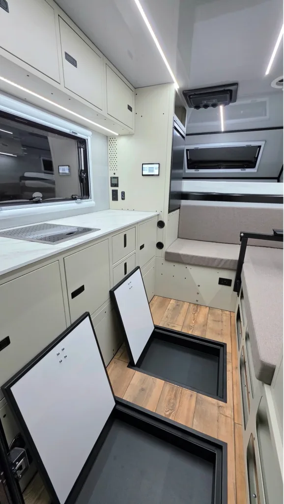 Modern camper van interior featuring a kitchenette with a sink, overhead cabinets, and bench seating. Two storage compartments are open in the wooden floor, revealing ample hidden storage space. Large windows provide natural light.