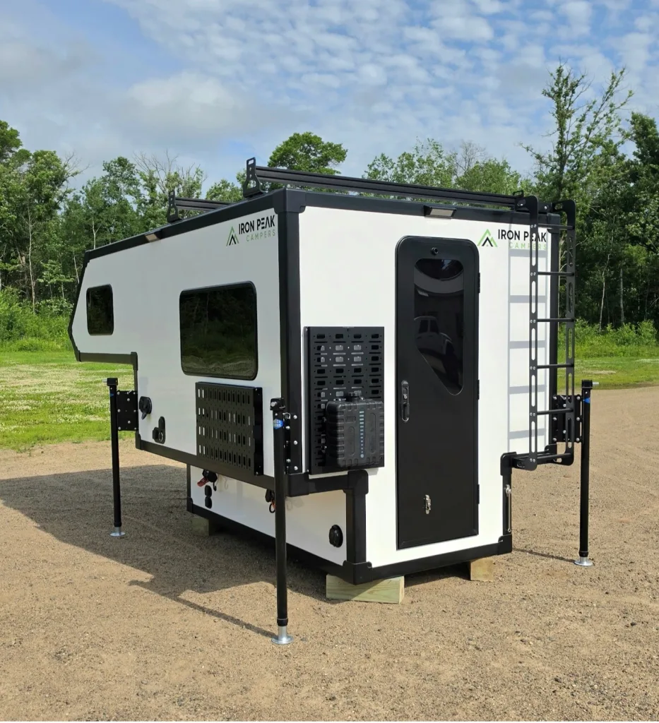 A white Iron Peak camper with black accents stands elevated on four support legs in a gravel area, surrounded by green grass and trees under a partly cloudy sky.