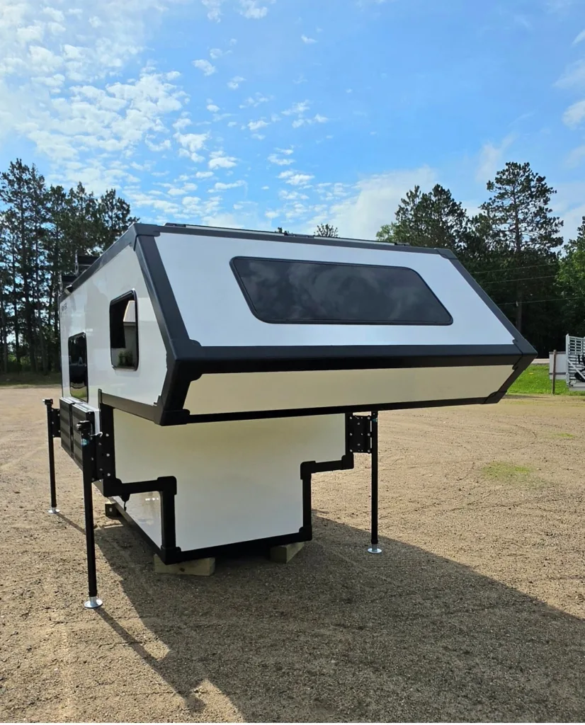 A modern white truck camper with black trim is elevated on four metal stilts in an open sandy area, surrounded by trees under a blue sky with scattered clouds.