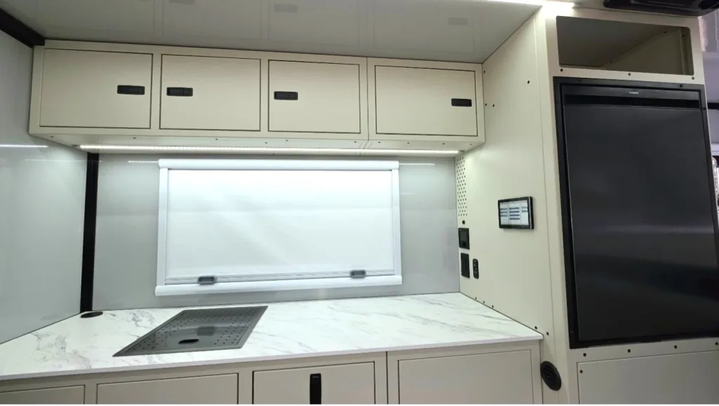 A modern RV kitchen with light cabinets, a marble-look countertop, an integrated stove, a window with a closed white shade, control panels on the wall, and a large black fridge on the right.