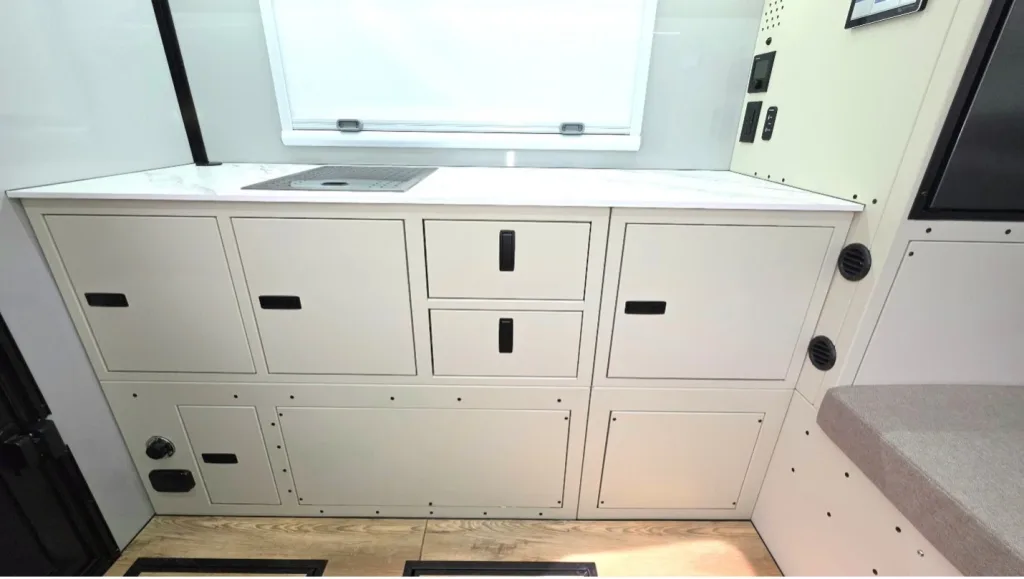 White cabinets and drawers with black handles and visible screws are built into a wall beneath a countertop with a sink. A window is above the counter, and part of a cushioned bench is visible on the right.