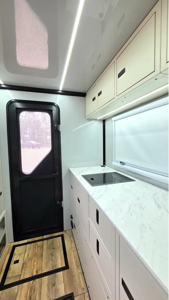 A modern RV kitchen with a black door, white cabinets, marble-style countertop, an induction cooktop, upper cupboards, and wood-patterned flooring. Natural light comes in through a large window with a shade.