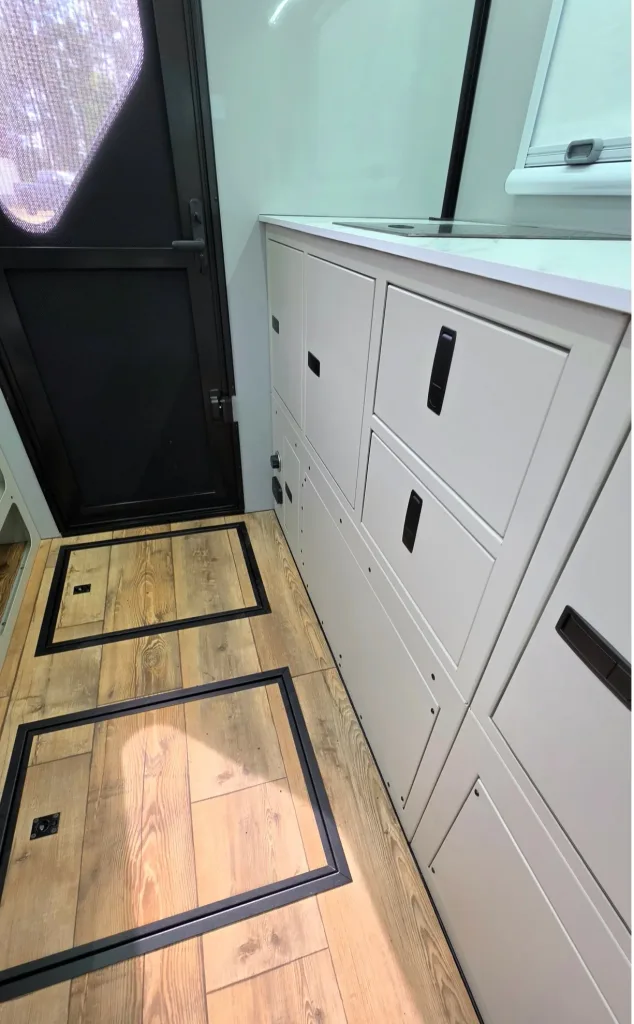Interior of a mobile workspace with white cabinets, a marble countertop, and a dark entry door on the left; wooden floor with black trim.