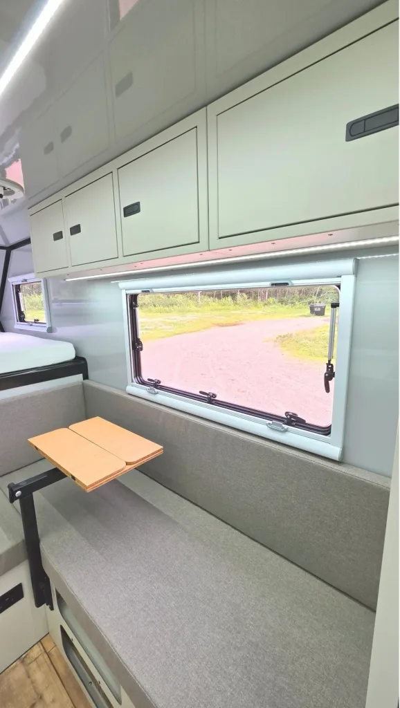 Interior of a camper van living space with grey cushioned bench seating, a small wooden table, white overhead cabinets, and a large side window.