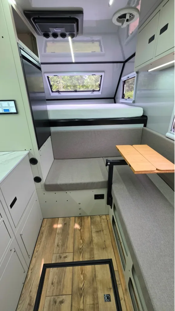 Interior of a compact camper van with light wood flooring, a gray sofa, a foldable wooden table, white cabinetry, and windows letting in natural light. A bed platform is positioned above the sofa area.