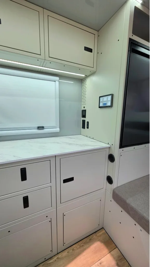 Compact, modern RV interior featuring white cabinetry with drawers and cupboards, a marble-look countertop, integrated control panel, and a window with a closed white shade above the workspace.