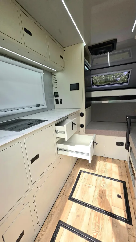 Modern camper van interior with light-colored cabinets, open drawers, wood floor with storage compartments, a small kitchen area, and a bench by a window with trees visible outside. The space looks clean and minimalistic.