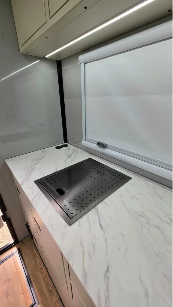 A modern kitchen countertop with a marble-like surface features a built-in sink covered by a dark perforated cover, white cabinets below, and a window with a closed white blind above.