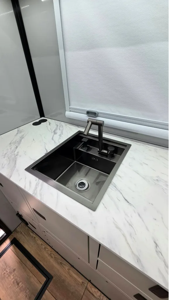 A modern stainless steel sink with a sleek faucet is set into a white marble countertop in a bright, minimalist kitchen. A window with a white shade is above the sink, and wood flooring is visible below.