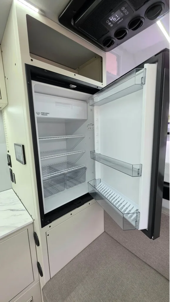 An open, empty refrigerator with glass shelves, a small freezer compartment at the top, and two clear drawers at the bottom; door shelves are visible, and the fridge is built into a modern white kitchen.
