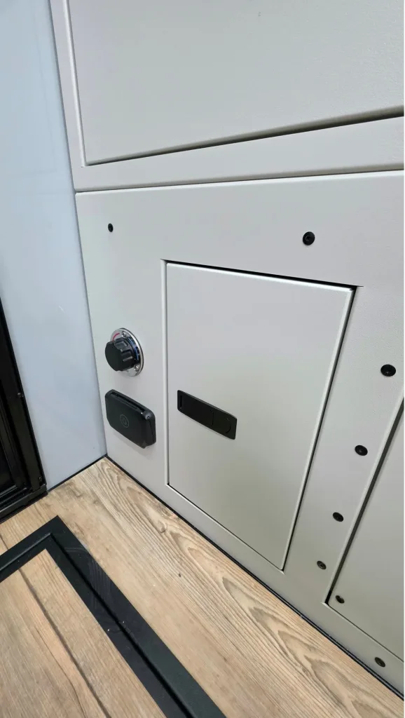 Close-up of a white metal cabinet door with a black latch, next to a wall with two black and silver control knobs or switches, above a wooden floor with a black strip along the edge.